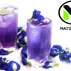 K&ouml;nnte beinhalten: Zwei hohe Gl&auml;ser gef&uuml;llt mit einem leuchtend violetten Getr&auml;nk, garniert mit Eis und einer Limettenscheibe. Die Getr&auml;nke sind von verstreuten blauen Schmetterlingserbsenbl&uuml;ten umgeben, was eine erfrischende und optisch ansprechende Pr&auml;sentation ergibt. Der Text "MAI'S FAMILY" ist enthalten.