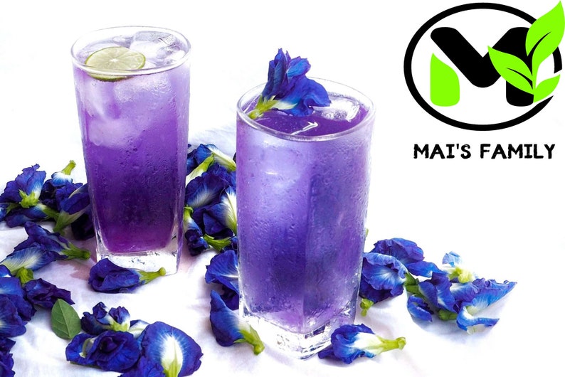 K&ouml;nnte beinhalten: Zwei hohe Gl&auml;ser gef&uuml;llt mit einem leuchtend violetten Getr&auml;nk, garniert mit Eis und einer Limettenscheibe. Die Getr&auml;nke sind von verstreuten blauen Schmetterlingserbsenbl&uuml;ten umgeben, was eine erfrischende und optisch ansprechende Pr&auml;sentation ergibt. Der Text "MAI'S FAMILY" ist enthalten.