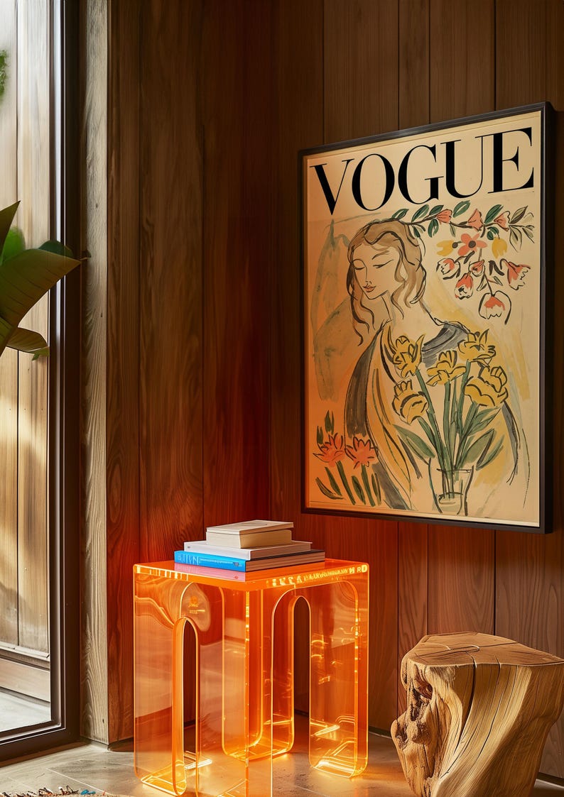 May include: A framed "VOGUE" magazine cover art print featuring a woman holding flowers. The artwork is displayed above a stack of books on a clear orange acrylic side table. A wooden stump is next to the table.