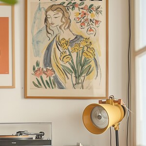May include: A framed Vogue magazine cover art print featuring a woman with flowers. The print has a wooden frame and is displayed on a white wall. A yellow lamp and stereo equipment are in the foreground.