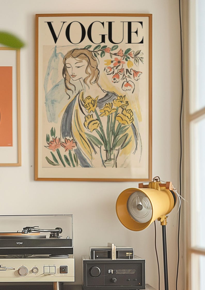 May include: A framed Vogue magazine cover art print featuring a woman with flowers. The print has a wooden frame and is displayed on a white wall. A yellow lamp and stereo equipment are in the foreground.
