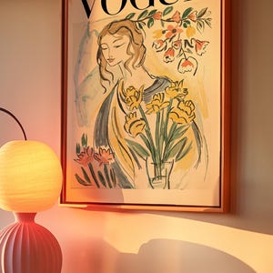 May include: Framed Vogue magazine cover art featuring a woman with flowers. The artwork has a vintage aesthetic with soft colors and a warm, inviting glow. The frame is a light brown color. A pink lamp and orange vase are in the foreground.