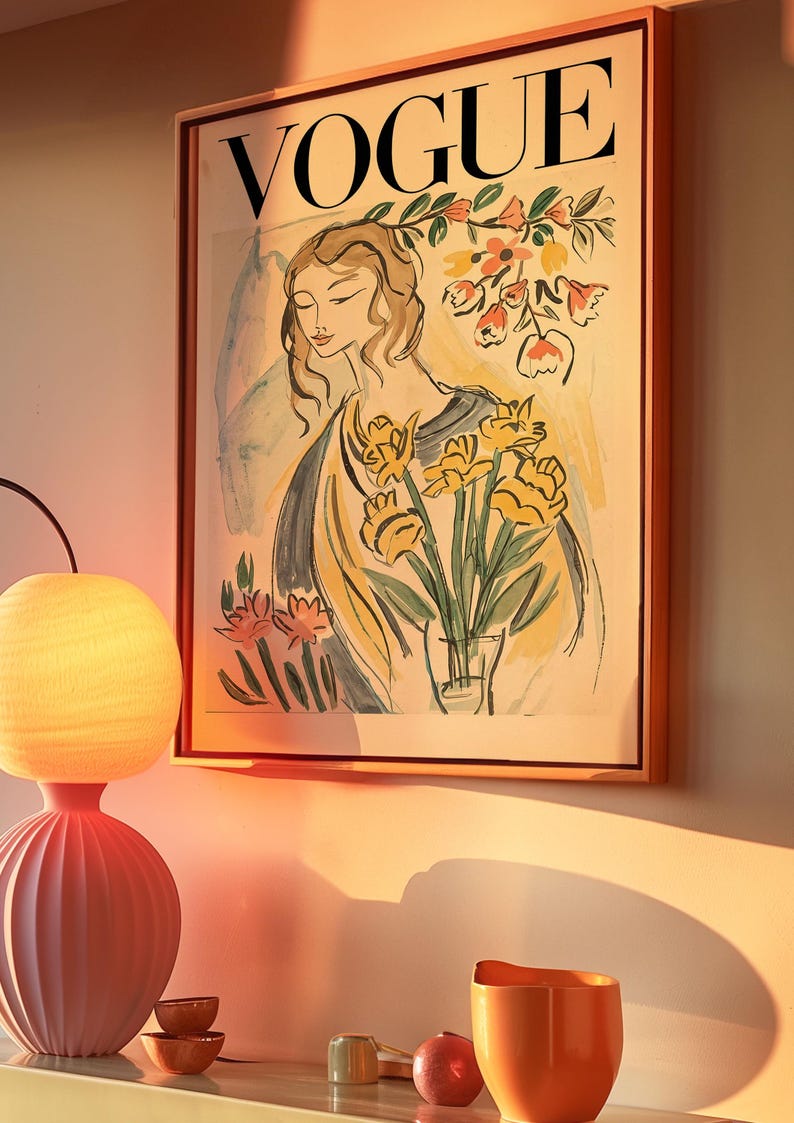 May include: Framed Vogue magazine cover art featuring a woman with flowers. The artwork has a vintage aesthetic with soft colors and a warm, inviting glow. The frame is a light brown color. A pink lamp and orange vase are in the foreground.