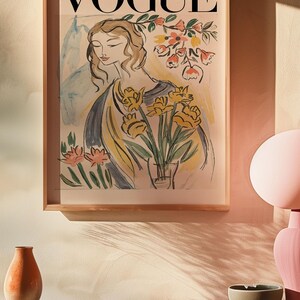 May include: A framed "VOGUE" magazine cover art print. The artwork features a watercolor illustration of a woman holding yellow flowers in a vase. The print is in a light wood frame, displayed on a shelf with decorative items.