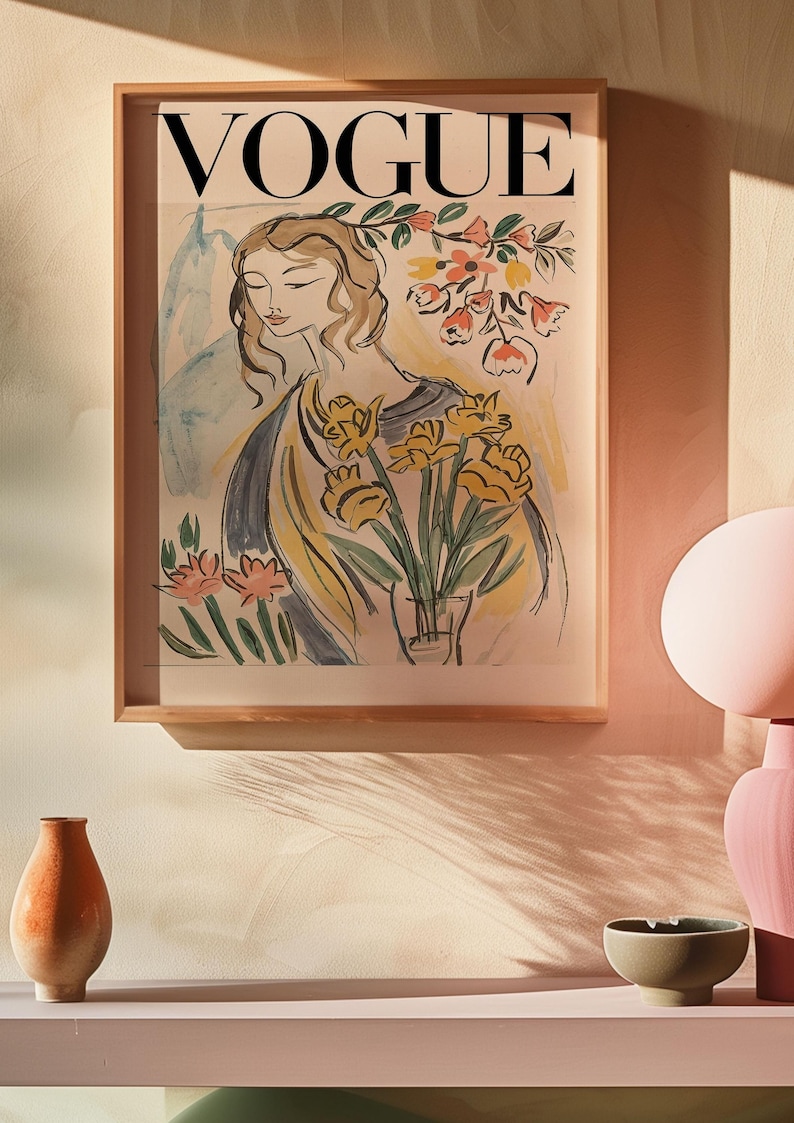 May include: A framed "VOGUE" magazine cover art print. The artwork features a watercolor illustration of a woman holding yellow flowers in a vase. The print is in a light wood frame, displayed on a shelf with decorative items.