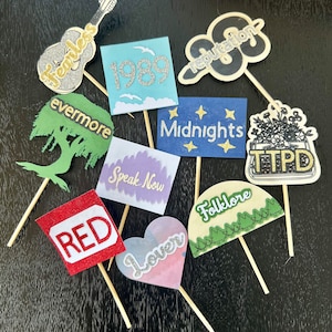 May include: A collection of ten colorful paper cupcake toppers featuring the names of Taylor Swift's albums, including "Fearless", "1989", "Reputation", "evermore", "Speak Now", "Midnights", "Taylor Swift", "Red", "Lover", and "Folklore".