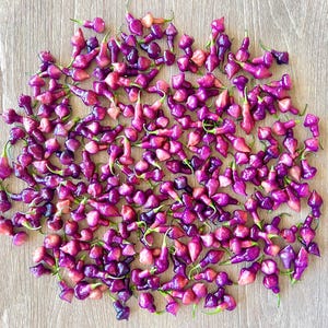 May include: A close-up of a large group of small, purple peppers. The peppers are arranged in a circular pattern on a light brown wooden surface.