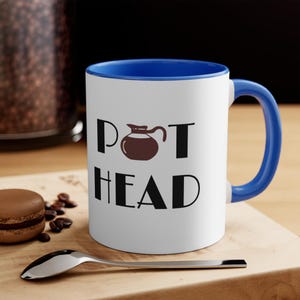 May include: White ceramic coffee mug with a blue rim and handle. The mug has a black graphic of a coffee pot and the text "POT HEAD".