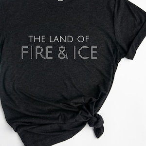 Op de afbeelding: Een donkergrijs T-shirt met de tekst "THE LAND OF FIRE & ICE" in wit gedrukt op de voorkant.