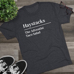 Peut inclure: T-shirt gris foncé avec le texte blanc "Haystacks The Adventist Taco Salad". Le t-shirt est en matière douce et est présenté sur une surface en bois avec des baskets noires et une petite plante.
