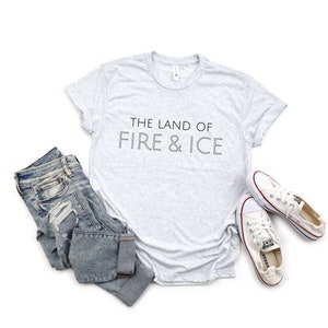 Op de afbeelding: Een grijs t-shirt met de tekst "THE LAND OF FIRE & ICE" in zwart gedrukt.