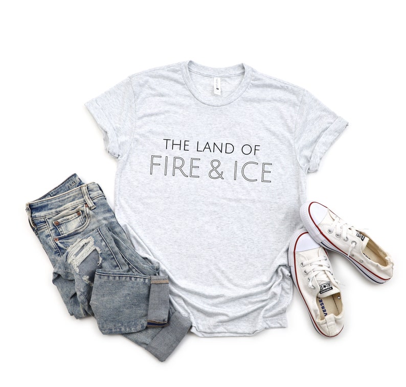 Op de afbeelding: Een grijs t-shirt met de tekst "THE LAND OF FIRE & ICE" in zwart gedrukt.