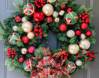 May include: A festive Christmas wreath with green faux pine needles, red berries, and an assortment of ornaments. The ornaments include gold, red, and green with gold accents. A plaid bow adds a decorative touch.