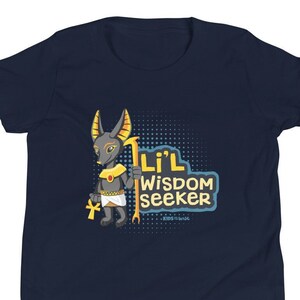 Può includere: T-shirt blu navy con un personaggio dei cartoni animati di Anubi che tiene in mano un bastone e un ankh. Il testo "Li'l Wisdom Seeker" è visualizzato in un carattere stilizzato. Il design incorpora accenti gialli, bianchi e neri.