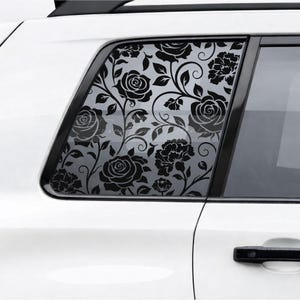 May include: A black floral decal applied to the rear side window of a white vehicle. The design features a repeating pattern of roses and swirling vines that cover the glass surface, creating a high-contrast graphic against the white car body.