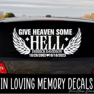 May include: A white vinyl car decal featuring the text GIVE HEAVEN SOME HELL, the name DERRICK DAVIDSON, and the dates 10/28/2002 heart 10/18/2023. The design includes two large angel wings flanking the word HELL and two small four-pointed stars.
