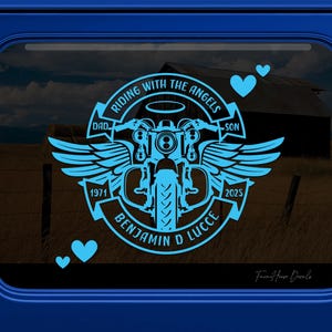 May include: A light blue vinyl decal featuring a motorcycle with wings and a halo. The text reads RIDING WITH THE ANGELS, DAD, SON, 1971, 2025, and BENJAMIN D LUCCE. Three small hearts are positioned around the central circular emblem design.