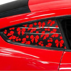 May include: Red car window louvers with a black background and a repeating pattern of red bows and cherries. The louvers are installed on a red car, adding a decorative touch to the vehicle's exterior.