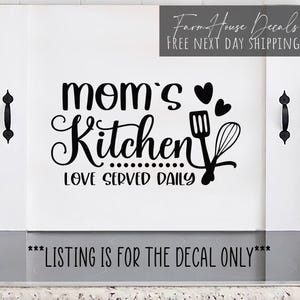 May include: A white rectangular stove cover board featuring black vinyl lettering that reads mom's Kitchen LOVE SERVED DAILY, accompanied by graphics of a spatula, a whisk, and two hearts, with black metal handles attached to the side panels.