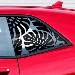 May include: A black vinyl decal featuring a repeating monstera leaf pattern applied to the rear side window of a bright red car. The decal covers the glass surface with large, stylized tropical foliage shapes, creating a bold contrast against the red.