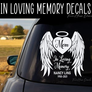 May include: A white vinyl car decal featuring a heart with angel wings and a halo above it. The heart contains the word Mom in script. Below the heart, the text reads In Loving Memory, NANCY LING, and the years 1950-2021 in white lettering.
