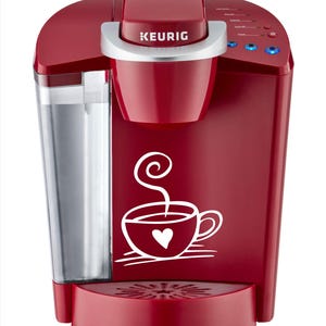 May include: A deep red Keurig coffee maker featuring a white graphic of a steaming cup with a heart on the front. The machine includes a clear water reservoir on the left side, a silver handle, and a control panel with blue indicator lights on top.