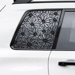 May include: A black floral decal applied to the rear side window of a white vehicle. The design features a repeating pattern of daisy-like flowers with detailed centers and petals, covering the entire glass surface of the car window panel.