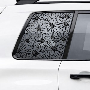 May include: A white car with a black floral design on the rear passenger window. The design features a repeating pattern of daisy-like flowers. The car door handle is black.