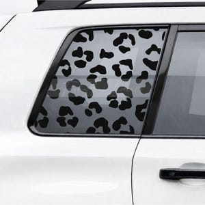 May include: A black leopard print decal applied to the rear side window of a white vehicle. The pattern consists of irregular, solid black spots distributed across the glass surface, creating a high-contrast graphic design on the car window.