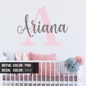 May include: A wall decal featuring a large light pink capital letter A with the name Ariana written in a gray cursive script font layered over the center. The design is displayed on a white wall above a white crib with pillows and pom-poms.