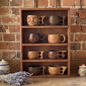 May include: A wooden shelf displays various handcrafted wooden mugs and cups. The mugs vary in shape and design, some with handles and others with unique carved details. The shelf is set against a rustic brick wall, with a wooden countertop.