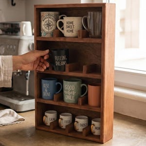 May include: A wooden four-tier mug display rack featuring various ceramic mugs with text including Home Sweet Home, Fueling Dreams, Rise & Shine, Good Vibes Only, Calm, Focus, Energy, and Mom. The rack has individual dividers for each mug.