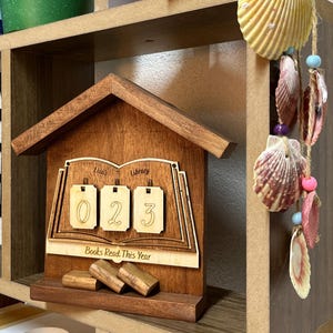 May include: A wooden bookshelf with books, a green potted plant, and a decorative wooden house-shaped book counter. The counter displays the numbers "023" and the text "Books Read This Year". Seashells and other decorative items are also present.