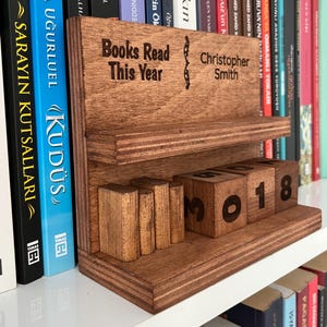 May include: A wooden book tracker stand with a shelf and three wooden blocks displaying the number 3018. The stand features the engraved text "Books Read This Year" and "Christopher Smith" on the back panel, positioned among various books.