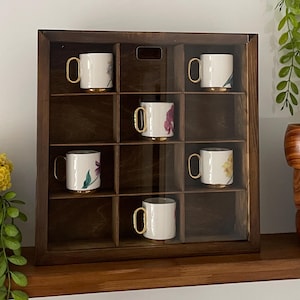 May include: A dark brown wooden display case with twelve square compartments, containing six white ceramic mugs. Each mug features a floral design, a gold-colored handle, and a gold-colored base. The case has a clear front panel.
