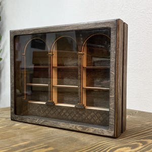 Könnte beinhalten: Eine Holzvitrine mit dunkelbraunem Finish. Die Vitrine hat drei gewölbte Glasscheiben, die jeweils ein kleines Regal umrahmen. Das Innere ist durch das Glas sichtbar und zeigt die Regale und einen dekorativen Sockel. Die Vitrine ist für die Aufstellung auf einer ebenen Fläche konzipiert.