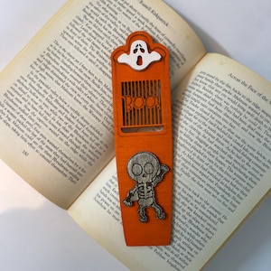 Könnte beinhalten: Oranger Halloween-Lesezeichen mit einem Geisterausschnitt oben und einem Skelett unten. Das Wort "BOO" ist in der Mitte ausgeschnitten. Das Lesezeichen liegt auf den Seiten eines Buches.