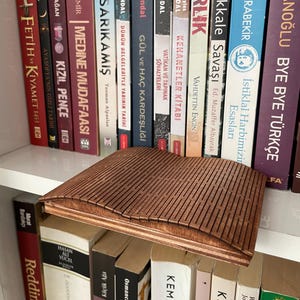 Könnte beinhalten: Ein brauner Buchhalter aus Holz mit Lamellendesign, der auf einem Regal mit Büchern liegt. Der Halter hat eine warme braune Farbe und scheint aus geschichtetem Holz gefertigt zu sein. Die Bücher haben verschiedene Titel und Farben.
