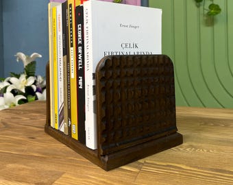 May include: A dark brown wooden bookend with a decorative carved pattern. The bookend is holding up a stack of books, including "1984" by George Orwell and "Çelik Fırtınalarında" by Ernst Jünger.