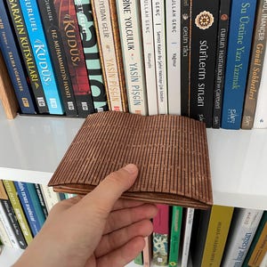 Könnte beinhalten: Ein brauner Buchhalter aus Holz mit geometrischem Design wird vor einem Bücherregal mit Büchern gehalten. Der Buchhalter besteht aus dünnen, parallelen Holzlatten. Die Bücher haben verschiedene Farben und Titel.