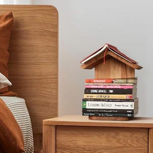 Könnte beinhalten: Ein Nachttisch aus hellem Holz mit einer Schublade und einem Regal. Ein Stapel Bücher wird von einem Miniatur-Holzhaus gekrönt. Der Bettrahmen und die Bettwäsche sind im Hintergrund zu sehen, mit einem terracottafarbenen Bettbezug.