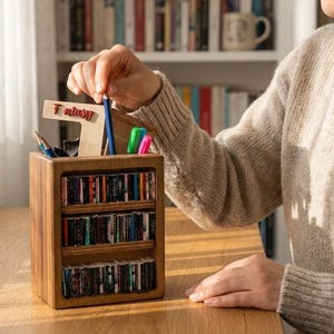 Puede incluir: Un organizador de escritorio de madera con tres estantes horizontales llenos de lomos de libros en miniatura y un compartimento superior para bolígrafos. Un letrero de madera con la palabra Fantasy está fijado arriba. Acabado en madera natural.