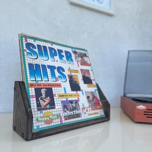 Puede incluir: Un soporte de madera oscura para discos que muestra una portada de álbum vintage "Super Hits". El álbum presenta imágenes de varios artistas y el texto "Happy Music". El soporte está sobre una superficie blanca.