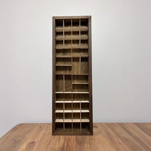 Puede incluir: Una estantería de madera rectangular y alta con múltiples compartimentos pequeños. La estantería es de madera marrón oscuro con veta natural, y los compartimentos están dispuestos en un patrón de rejilla. Diseñada para exhibir objetos pequeños.