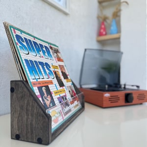 May include: A dark wooden record holder displaying a vintage "Super Hits" album. The album cover has a grid pattern and artist photos. A retro-style orange record player is in the background.
