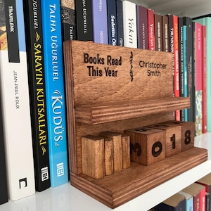 May include: A wooden bookshelf organizer with the text "Books Read This Year" and "Christopher Smith" engraved on it. The organizer has a shelf and slots for wooden blocks with the year 2018. Books are visible in the background.
