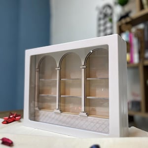Op de afbeelding: Een witte schaduwdoos met drie boogvormige openingen, elk met een plank. De binnenkant heeft een lichtgekleurde, patroonvloer. De doos is rechthoekig met een doorzichtig voorpaneel. De achtergrond is wazig, met hints van blauw en een boekenplank.