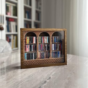 May include: A rectangular wooden shadow box frame containing a miniature three-arched bookshelf display. The interior features three levels of tiny, colorful book spines arranged on shelves, with a decorative lattice pattern along the bottom base.