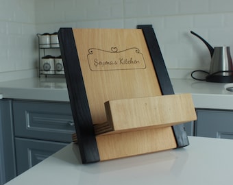 May include: A wooden cookbook stand featuring a light natural wood finish with contrasting black side supports and a horizontal ledge. The centre panel is engraved with a decorative border, a small heart icon, and the text "Şeyma's Kitchen".