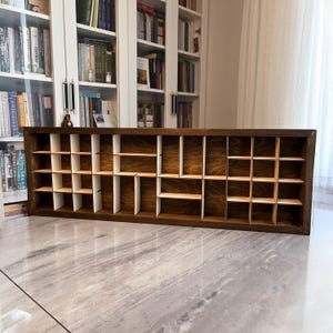 Puede incluir: Estantería rectangular de madera marrón oscuro con múltiples compartimentos pequeños. La estantería tiene un acabado de veta de madera natural. Los compartimentos están divididos por separadores verticales y horizontales, creando un patrón de cuadrícula. Ideal para organizar y exhibir objetos pequeños.